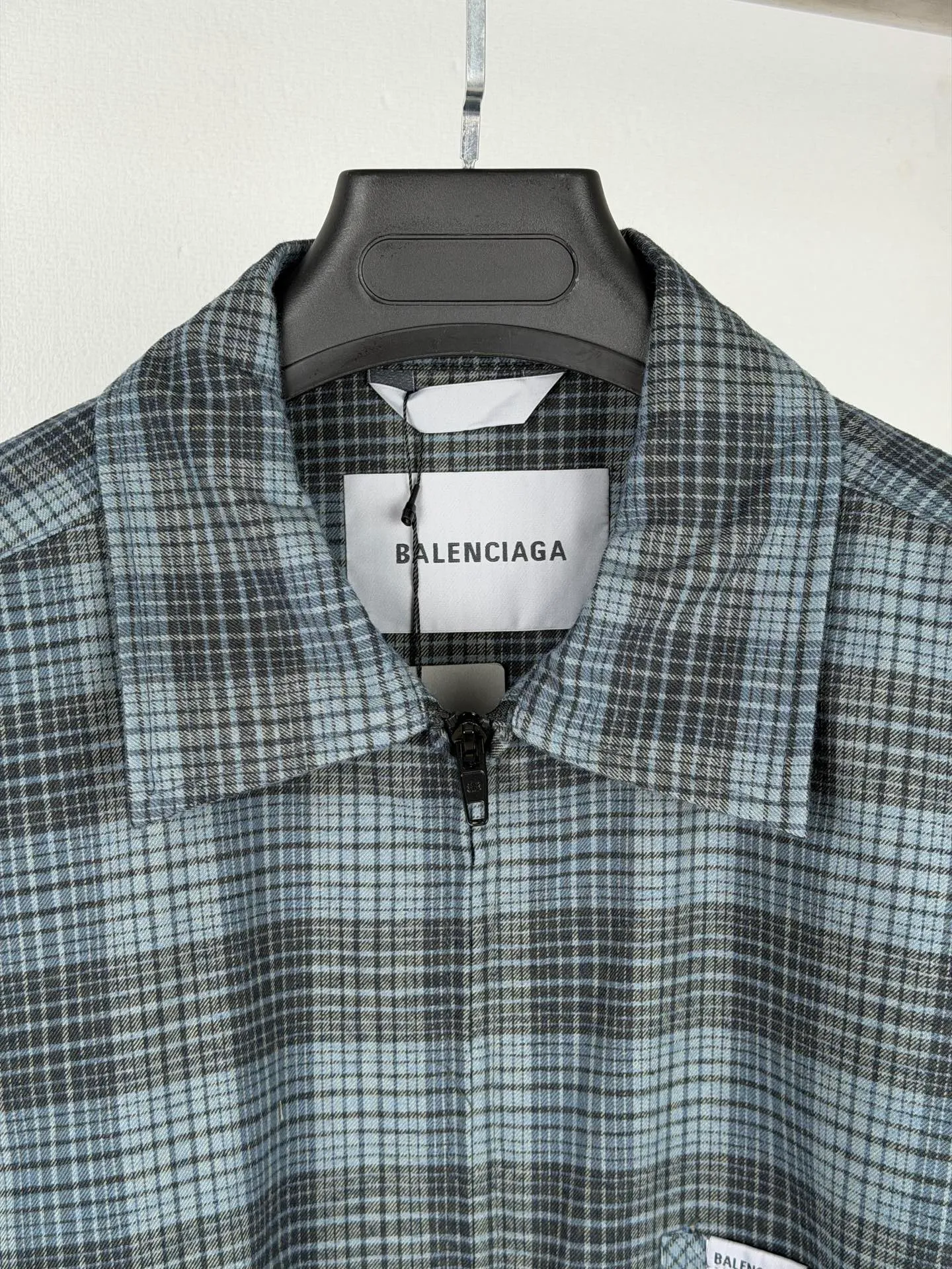 BALENCIAGA/balenciaga blue and gray plaid zipper pocket logo quilted jacket combines a three-layer structure of plaid woven fabric, filling cotton, and lining, and integrates plaid alignment technology and quilted garment production standards. There are multiple process difficulties in the whole process. The surface layer of the body is made of blue-gray Retro plaid fabric, and the cutting machine production strictly follows the global grid standards. The arrangement direction of the cutting pieces is fixed and cannot be changed at will. The horizontal and vertical lines of shoulder seams, side seams, and zipper placket joints must be consistent and coherent. The left chest patch pocket is cut independently, and the bag check pattern must be aligned with the front reference pattern. Slight deviations will destroy the visual integrity and directly increase fabric loss and cutting time. The most difficult part of the style is the three-layer composite sewing. The outer plaid fabric, filling cotton, and lining fabric have different physical properties, and the preshrinking processing parameters need to be adjusted separately. Pressure and temperature control are crucial during lamination operations. Once the parameters are out of balance, it can easily cause fabric stretch and distortion, plaid dislocation, and wrinkles and bulges in garments. At the same time, it is necessary to reasonably plan the quilting line to avoid the visual beauty of the grid pattern from being cut by the lines on the basis of firmly fixing the cotton layer and preventing cotton from running away. The placket adopts a centered zipper structure. It is difficult to sew the zipper tape and the quilted panels. It is required that the width of the left and right plackets be equal, the zipper routing is straight and smooth, and there is no lag in opening and closing. When the shirt lapel is filled with cotton, the thickness of the cotton needs to be controlled to prevent the collar from becoming bloated and unnaturally folded. The metal nameplate of the chest pocket is sewn at fixed points to avoid pulling the fabric during the operation and ensure that the plaid pattern is smooth and not deformed. The quilted material increases the overall thickness of the fabric. When sewing, you need to continuously adjust the presser foot pressure and stitch length to adapt to changes in sandwich thickness. The low-temperature and gentle ironing process is used for post-finishing to prevent the plaid from fading and the cotton layer from clumping due to high temperatures. From the precise alignment of cutting pieces, three-layer fabric pretreatment, composite quilting to garment finalization inspection, the processes are interlocking and the error tolerance rate is extremely low, testing the accuracy of plate making, equipment control and turning technology. Size:XS S M L
